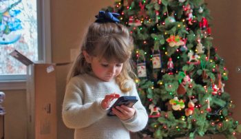 A girl using a smartphone next to a Christmas tree.